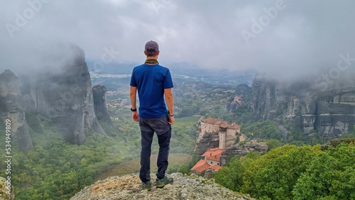 Man with scenic view of Holy Monastery of Rousanos appearing from fog, Kalambaka, Meteora, Thessaly, Greece, Europe. Mist atmosphere in dramatic landscape. Landmark build on unique rock formations photo
