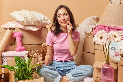 Dreamy pleased Asian woman sits crossed legs on floor around cardboard boxes relocates to new apartment dressed in casual t shirt jeans imagines design of her room surrounded by personal belongings
