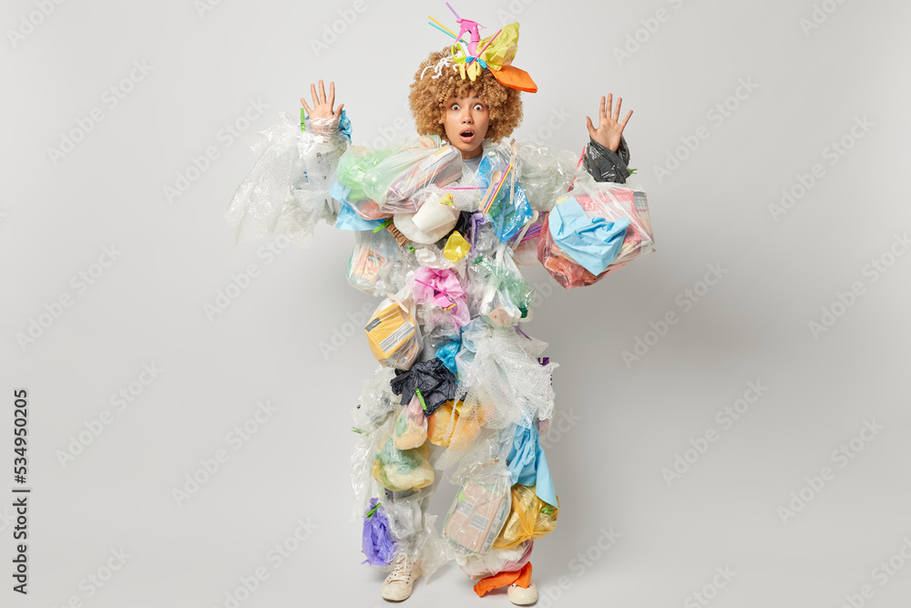 Scared frightened woman activist keeps hands raised stares at camera poses in rubbish costume takes care of environment cannot believe own eyes stands in full length against grey background.