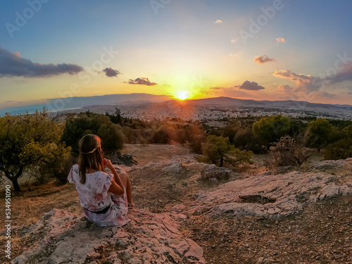 Tourist woman watching sunset over city of Athens seen from Filopappou Hill (hill of muses), Athens, Attica, Greece, Europe. Athens cityscape and Aegean sea. Beautiful sunset point with aerial view