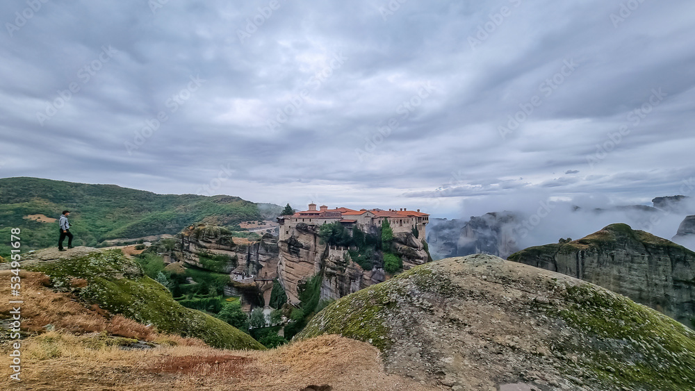 Man standing on cliff edge with panoramic view of Holy Monastery of Varlaam, Kalambaka, Meteora, Thessaly, Greece, Europe. Rock formations overgrown with moss, moody atmosphere. UNESCO World Heritage