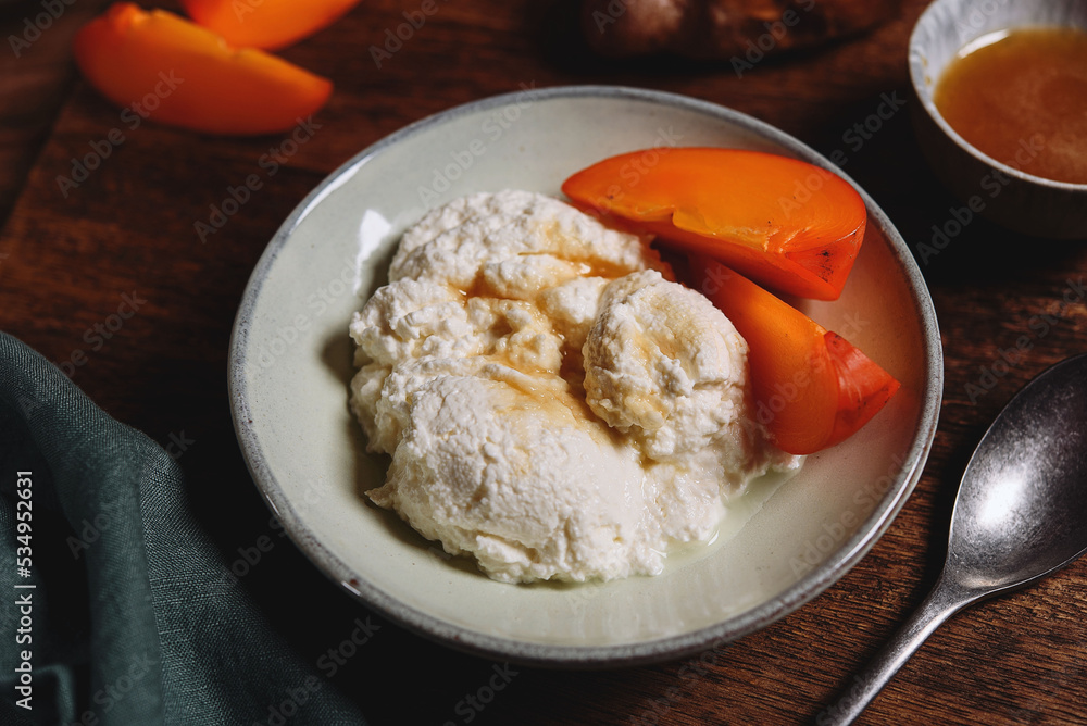 Homemade ricotta cheese or cheese curd served with fresh persimmon and honey and croissante on wooden background. Healthy breakfast. Haze effect. Selective focus Homemade ricotta cheese or cheese curd served with fresh persimmon and honey and croissante on wooden background. Healthy breakfast. Haze effect. Selective focus