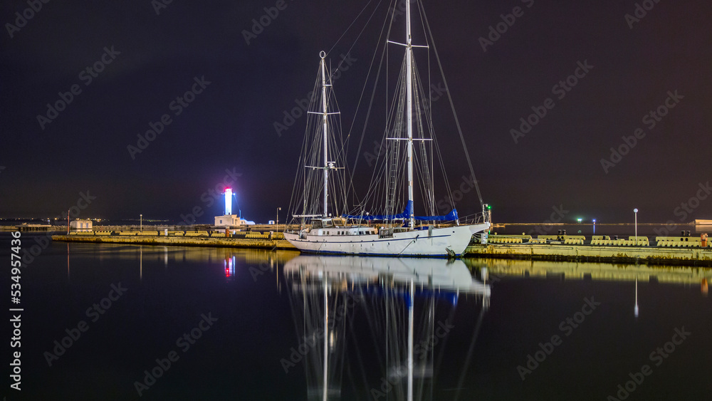 A white sailing ship with two masts stands in the port and is displayed in the water against the background of a lighthouse and the night sky