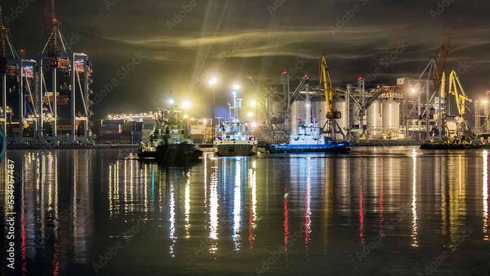A variety of small ships floating at night in the port against the background of port cranes, lanterns and containers
