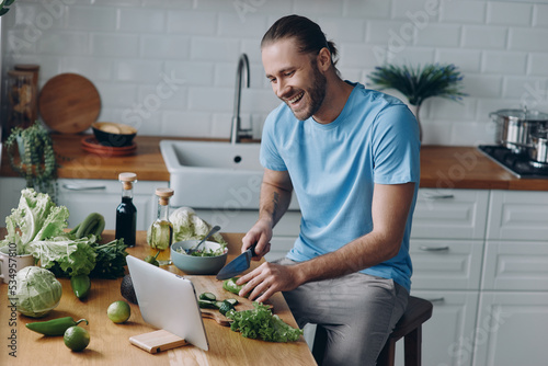 Happy young man looking at digital tablet while preparing food at the domestic kitchen photo