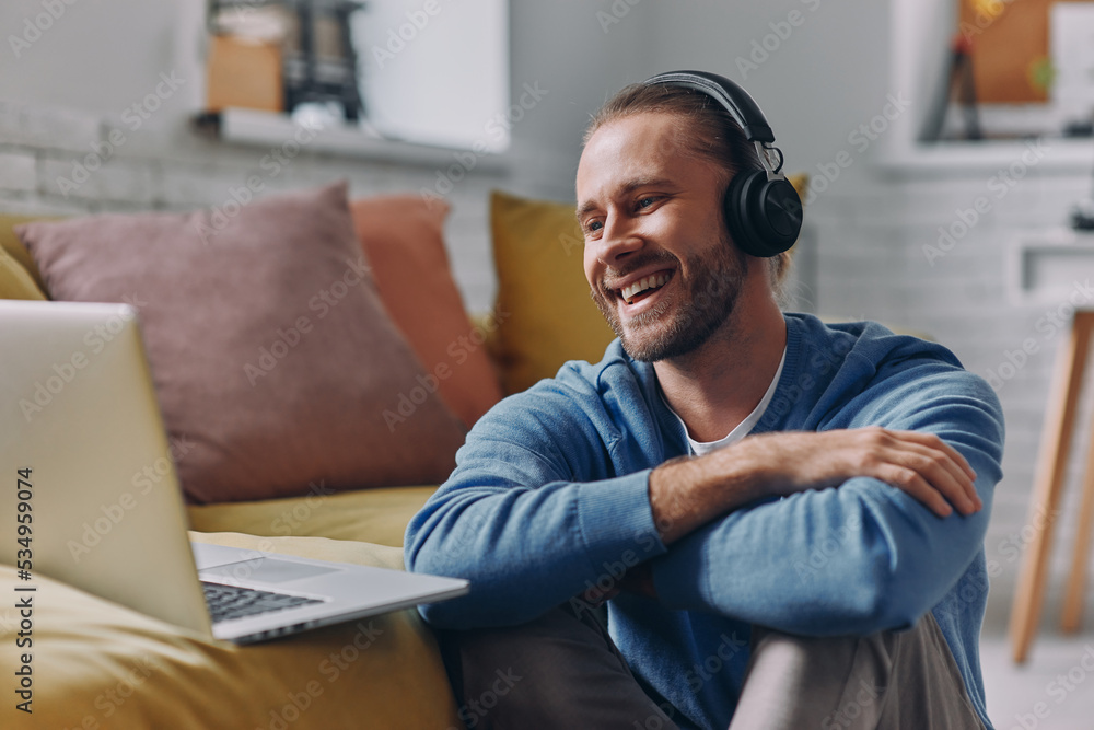 Handsome man in headphones looking at laptop and smiling while sitting on the floor at home Handsome man in headphones looking at laptop and smiling while sitting on the floor at home