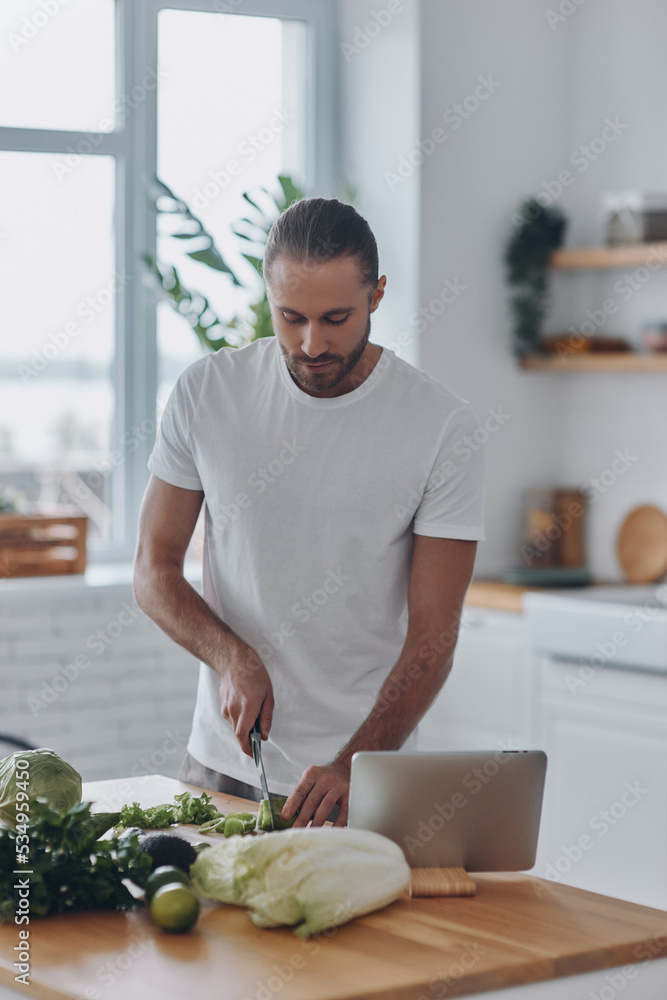Fototapeta premium Handsome young man cutting veggies while standing at the kitchen island at home