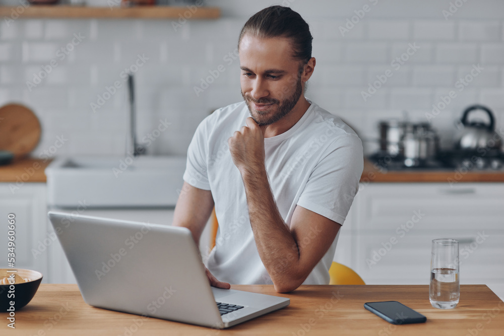 Handsome young man using laptop while working at the kitchen table at home Handsome young man using laptop while working at the kitchen table at home