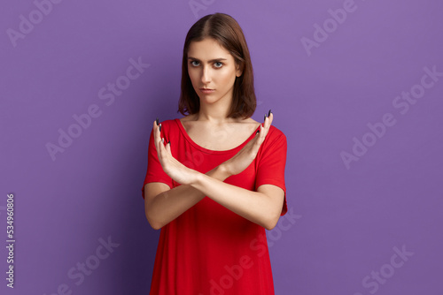 Access denial. It is forbidden. Protective young pretty brunette girl in red t-shirt show ban gesture with crossing hands. Looking at the camera. Posing over purple background.
