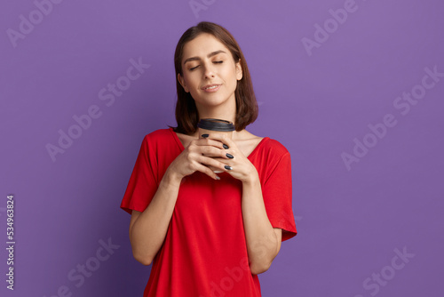 Coffee is a best drink. Satisfied young pretty brunette girl in red t-shirt holds disposable cup and close her eyes in pleasant mood. enjoys caffeine beverage aroma. Posing over purple wall.