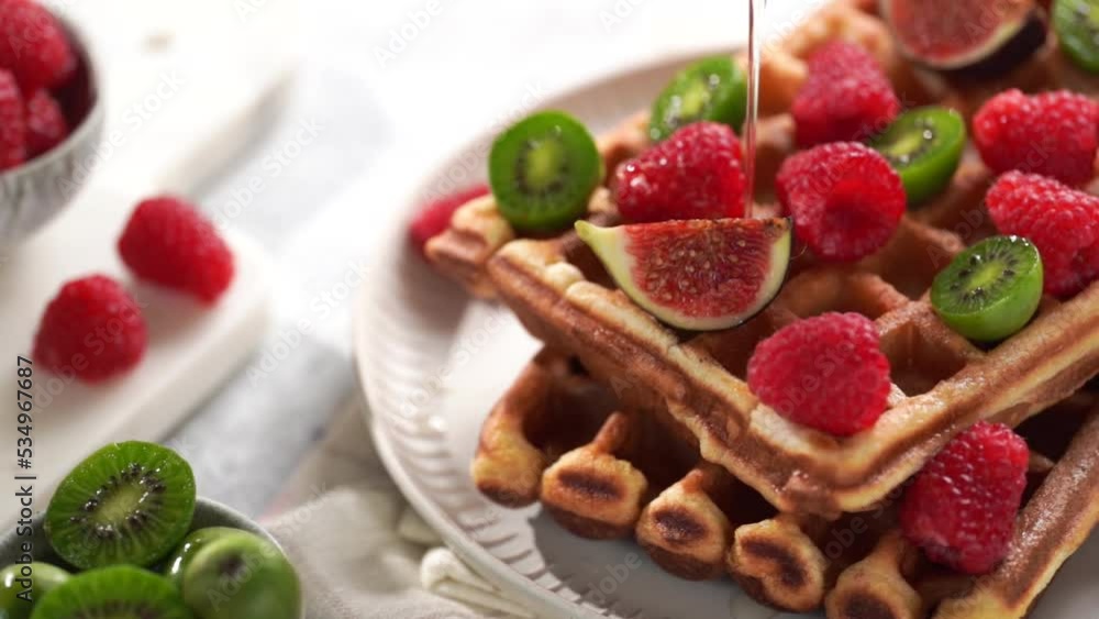 Pouring honey over fresh homemade sweet belgian waffles with powdered sugar with fresh raspberries, kiwi berries and figs on white plate Pouring honey over fresh homemade sweet belgian waffles with powdered sugar with fresh raspberries, kiwi berries and figs on white plate