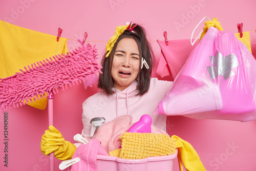 Dissatisfied crying woman does housework holds big rubbish bag and mop stands near basin full of laundry tired of housework wears hoodie and rubber gloves stands against washline cleans house photo