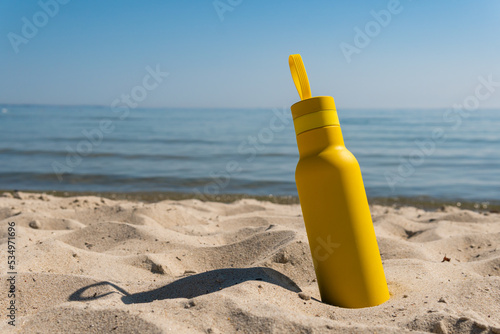A yellow bottle, thermos standing in the sand on a beach, ocean on sunny summer day. Symbol of summer, holidays and rest