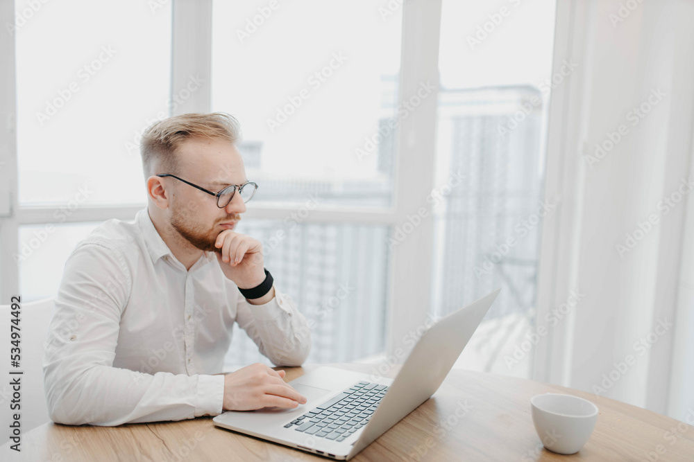 young european man in glasses works with a laptop at a table in a light coworking type office in a skyscraper. young european man in glasses works with a laptop at a table in a light coworking type office in a skyscraper.