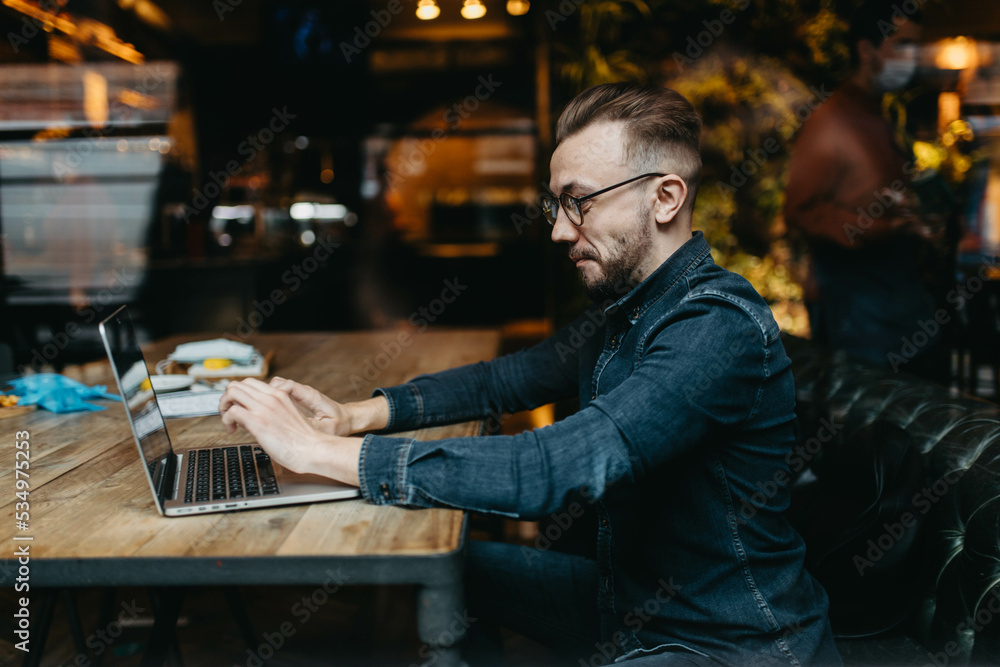 young male IT specialist with a beard and hairstyle works with a laptop in a cozy cafe. Online work on the Internet. young male IT specialist with a beard and hairstyle works with a laptop in a cozy cafe. Online work on the Internet.