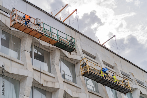 A worker in a cradle of a lifting mechanism on the roof restores cleans the marble wall of the city university close-up view from below.