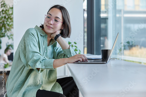Obraz na plátně Thoughtful Asian female freelancer works on remote project browses information in web site drinks coffee poses at windowsill in cozy cafe looks away with pensive expression