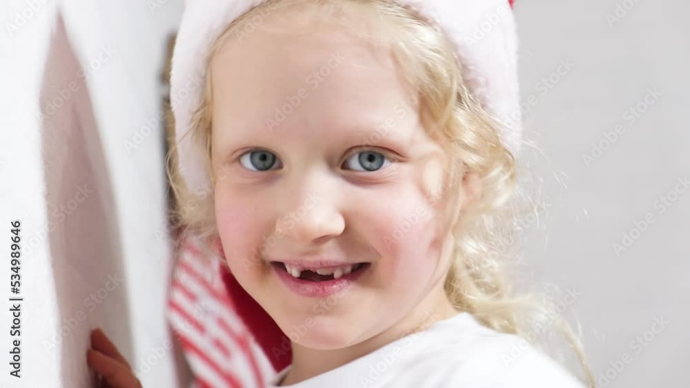 portrait of a cute curly girl with blond hair in a santa hat, the child has fun and makes faces on the eve of christmas girl's baby teeth fell out portrait of a cute curly girl with blond hair in a santa hat, the child has fun and makes faces on the eve of christmas girl's baby teeth fell out