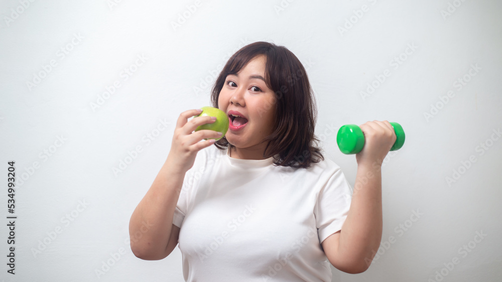 Chubby woman wearing white t-shirt holding dumbbells and apples poses for healthy exercise and eat fruit, health concept. Chubby woman wearing white t-shirt holding dumbbells and apples poses for healthy exercise and eat fruit, health concept.