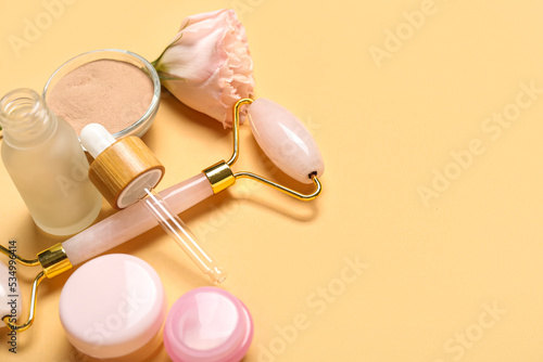Facial massage tool with cosmetic products and rose on beige background