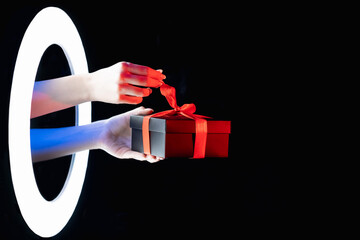 Holiday surprise. Black Friday sale. Giveaway promotion. Female hands untying gift box with red ribbon in LED ring color neon light isolated on dark night empty space background.