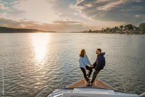 Young romantic couple standing on the prow of a boat on the river during sunset. River travel and honeymoon concept