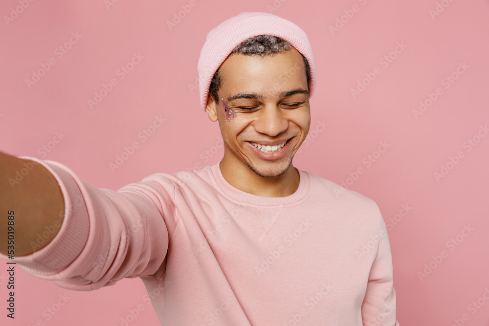 Close up young smiling happy gay man wear sweatshirt hat doing selfie shot pov on mobile cell phone isolated on plain pastel light pink color background studio portrait. Lifestyle lgbtq pride concept. Close up young smiling happy gay man wear sweatshirt hat doing selfie shot pov on mobile cell phone isolated on plain pastel light pink color background studio portrait. Lifestyle lgbtq pride concept.