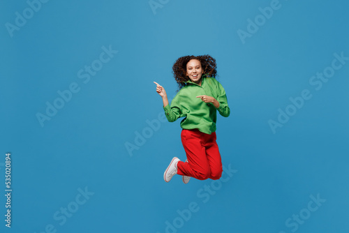 Full body young woman of African American ethnicity 20s she wear green shirt jump high point index finger aside on workspace area mock up isolated on plain blue background. People lifestyle concept.