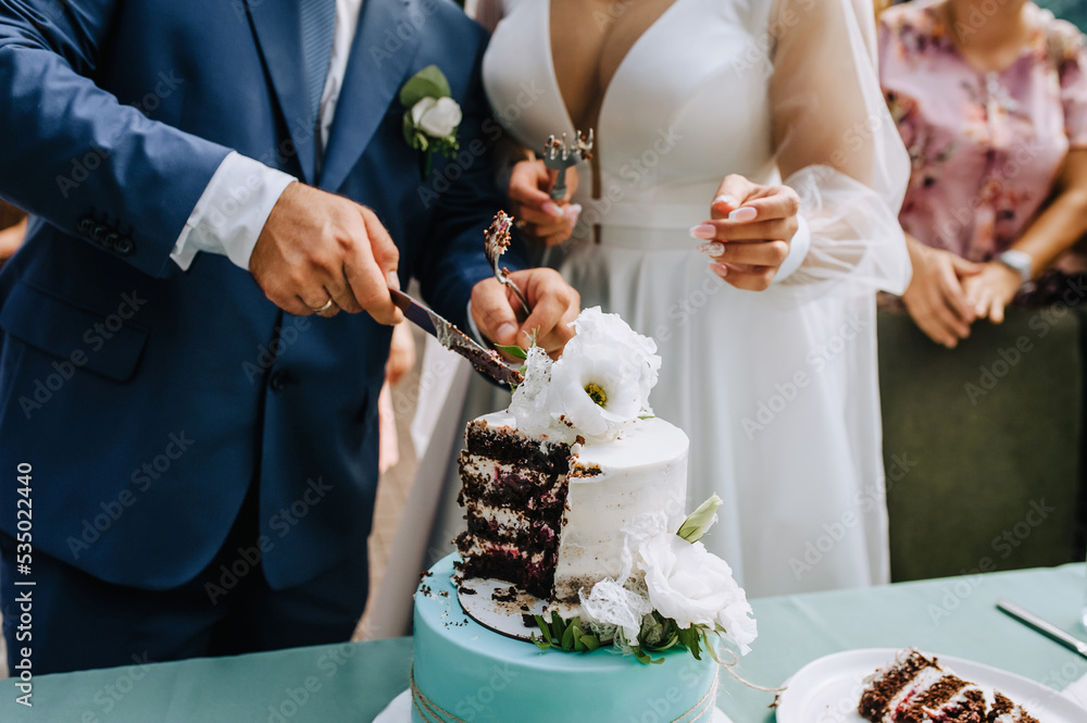Fototapeta premium The bride and groom cut the sweet, delicious wedding cake, which is on the table, into a piece with a knife at the ceremony.