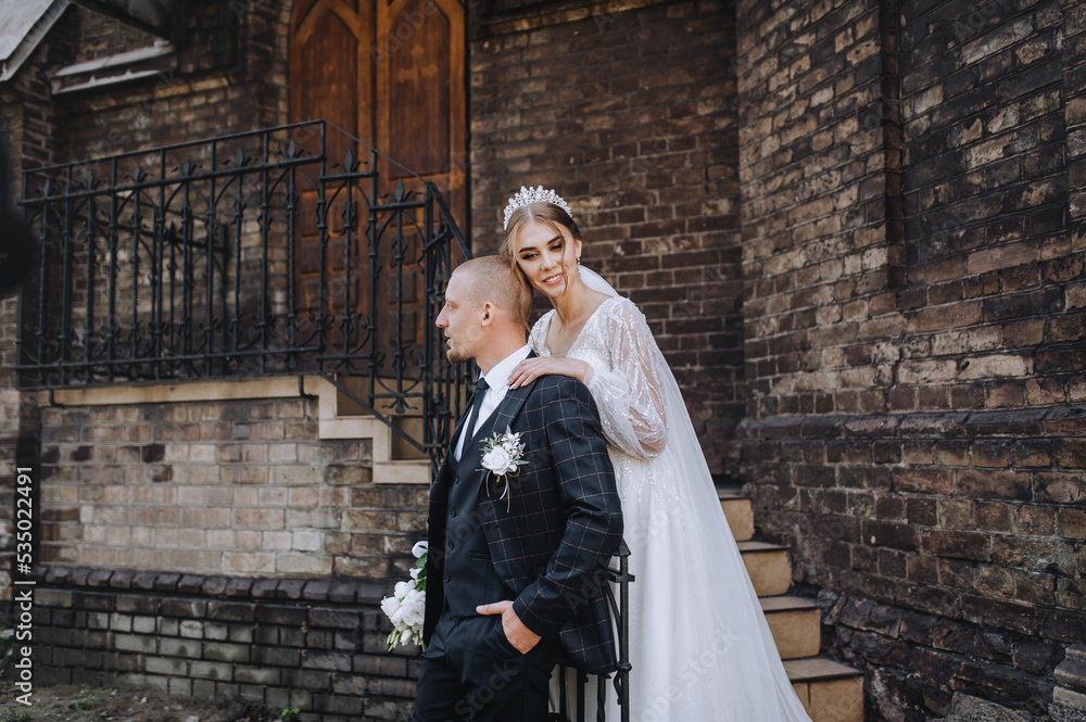 A young groom in a blue plaid suit and a cute bride in a white lace dress with a diadem, a bouquet are hugging, standing near the brick wall of the church, temple. Wedding photography, portrait.