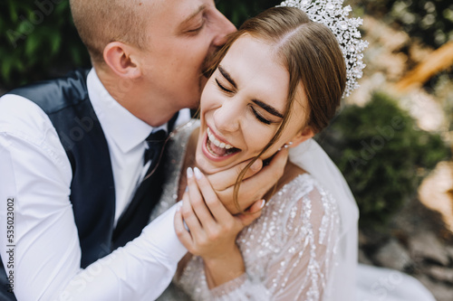 Wedding photo of cheerful, smiling, funny newlyweds. A stylish, young groom and a beautiful bride in a white dress with a bouquet in their hands, a diadem on their heads, sit in a park in nature.