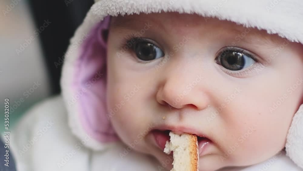 A beautiful caucasian baby girl in a fleece hat holds a crust of bread with her hand and nibbles with a toothless snail, side view close-up with depth of field and selective focus. A beautiful caucasian baby girl in a fleece hat holds a crust of bread with her hand and nibbles with a toothless snail, side view close-up with depth of field and selective focus.