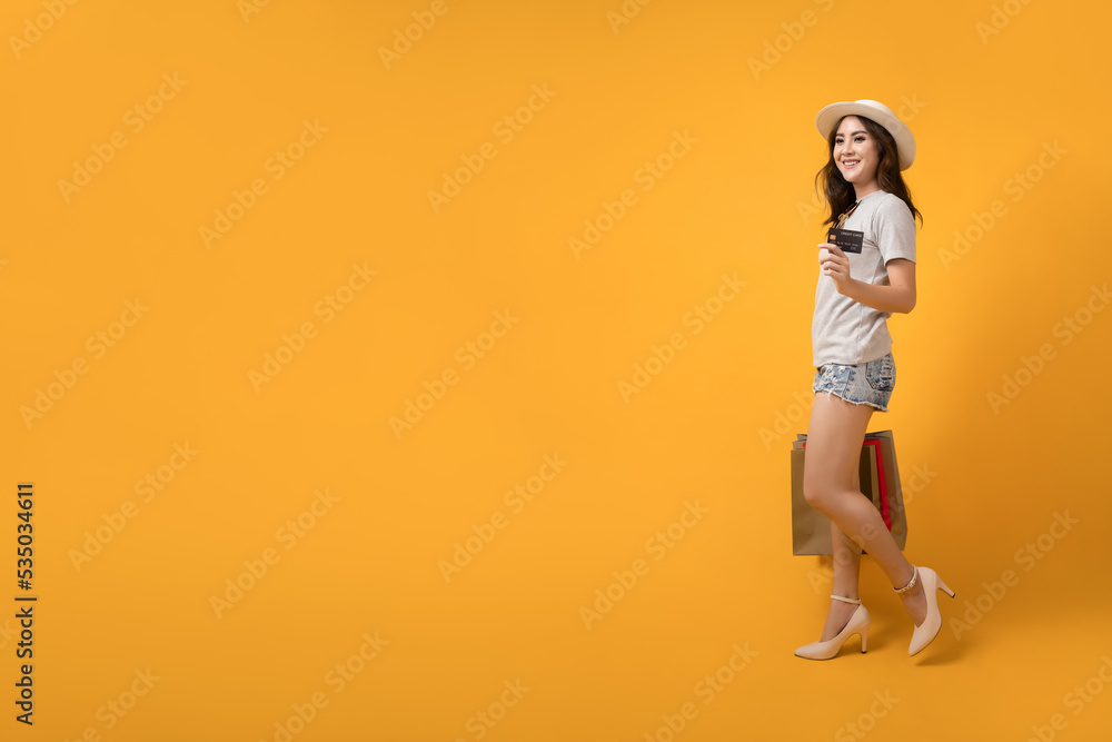 Beautiful Asian shopaholic woman smiles and happy while carrying shopping bags and credit card on yellow background. Summer sale, Mid-year sales concept.