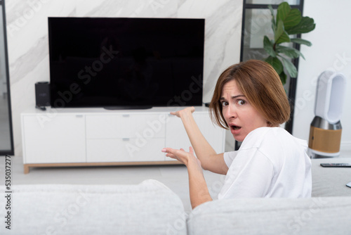 Shocked girl sitting on the couch, watching TV and pointing on screen. She demonstrating her disgusting. Black screen and free space on television concept