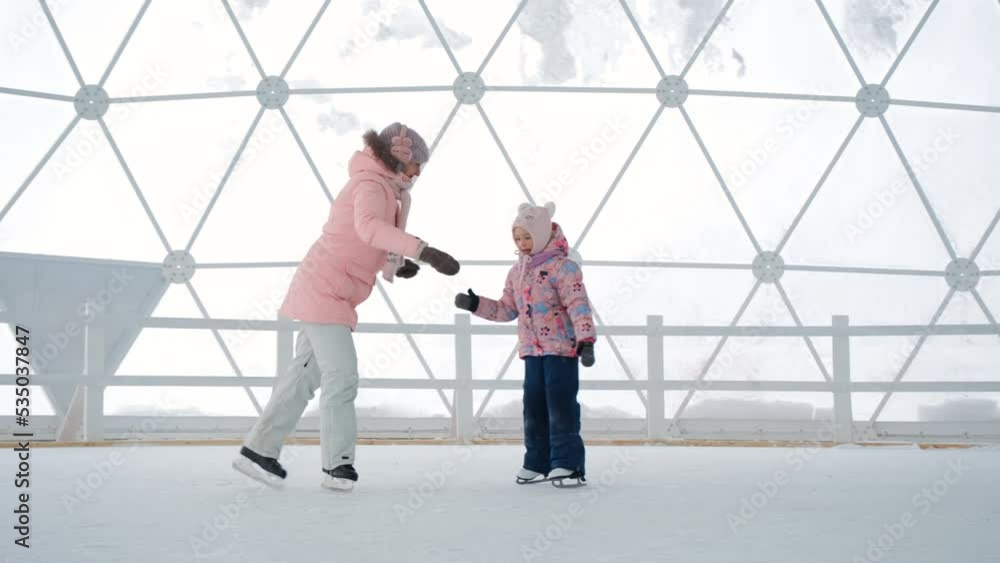 Young mother teaching her little daughter ice skating at indoor rick, slow motion. Winter activities concept Young mother teaching her little daughter ice skating at indoor rick, slow motion. Winter activities concept