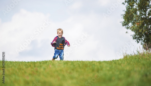 A little boy in a checkered shirt and blue pants is playing on top of a meadow near the horizon. Behind the child he saw the sky dotted with clouds. The child is blond and runs across the meadow.
