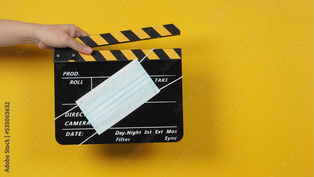  The hand is holding a black clapper board with a medical face mask on yellow background.