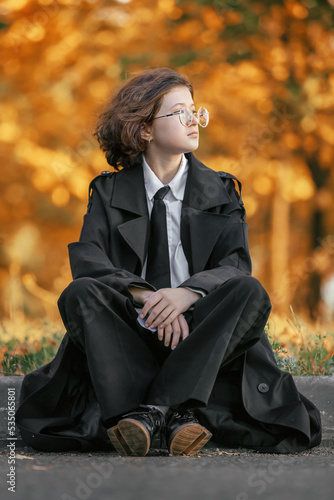 portrait of teenage girl with glasses in black raincoat sitting on asphalt road on curb on background of autumn forest, subculture of dark academia, autumn tinting, autumn background, October
