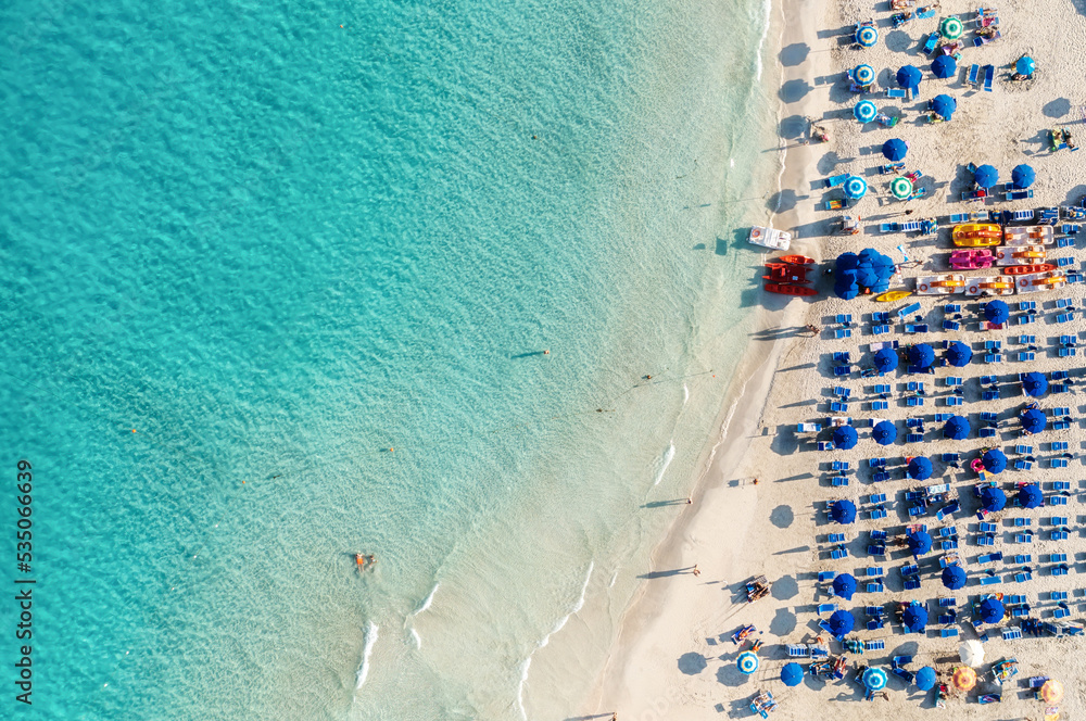 Top view of beautiful sandy popular beach La Pelosa with turquoise sea water and colorful blue umbrellas on the Sunset, Islands of Sardinia in Italy, aerial drone shot