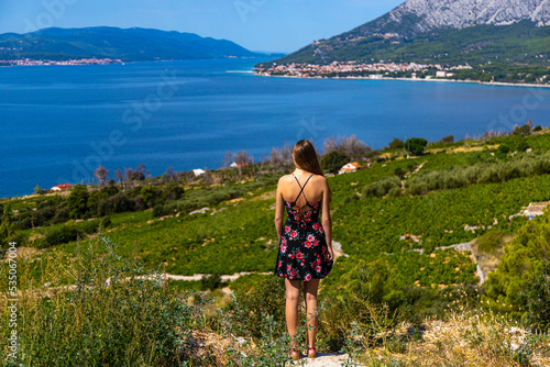 a beautiful long-haired girl in a black summer dress stands on top of a mountain overlooking a paradisiacal Croatian bay and the mighty green mountains in the background; vacation in croatia on the pe