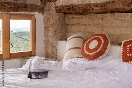 Interior habitación con ventana con vistas a la naturaleza, cama con sábanas blancas, ordenador encima de la cama, nordico. En la mesita libros. photo