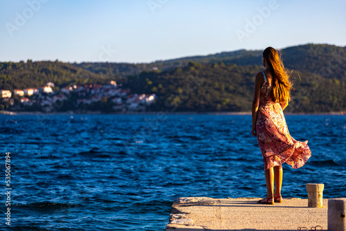 a beautiful long-haired girl in a long pink dress enjoys the sunset at the marina in the town of orebić, croatia, the peljesac peninsula and its paradise bays