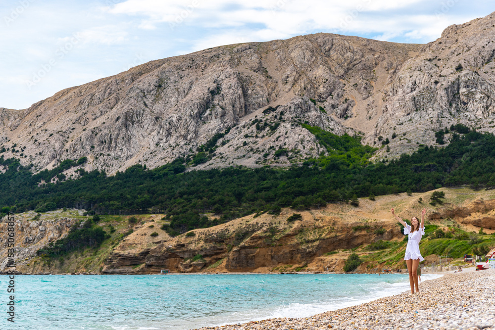 beautiful woman in a classic white dress enjoys a sunny day on a paradise beach in croatia, bay surrounded by mountains, krk island in the mediterranean sea