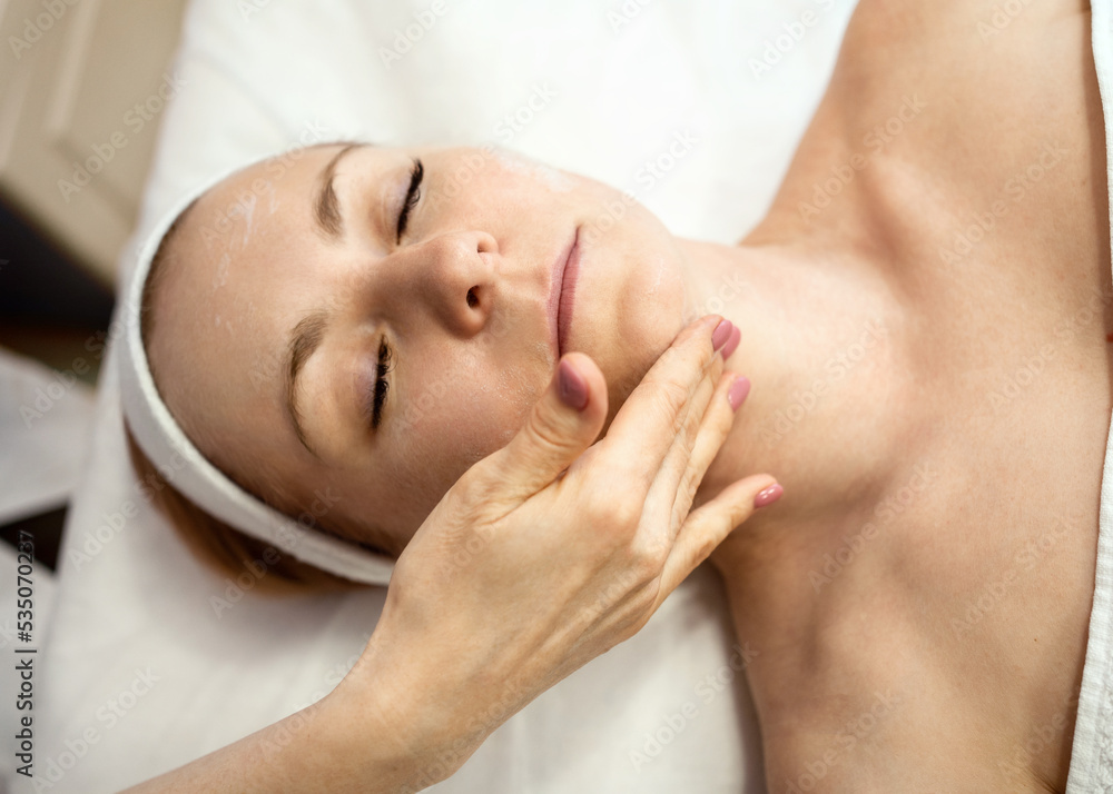 Hands of a massage therapist in a beauty salon performing a cleansing procedure. A young pretty woman with closed eyes lies relaxed on a massage table on a white bedspread and covered with a white