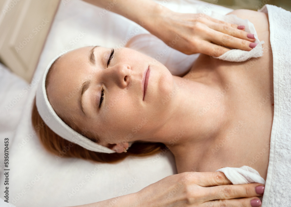 Hands of a massage therapist in a beauty salon performing a cleansing procedure. A young pretty woman with closed eyes lies relaxed on a massage table on a white bedspread and covered with a white Hands of a massage therapist in a beauty salon performing a cleansing procedure. A young pretty woman with closed eyes lies relaxed on a massage table on a white bedspread and covered with a white