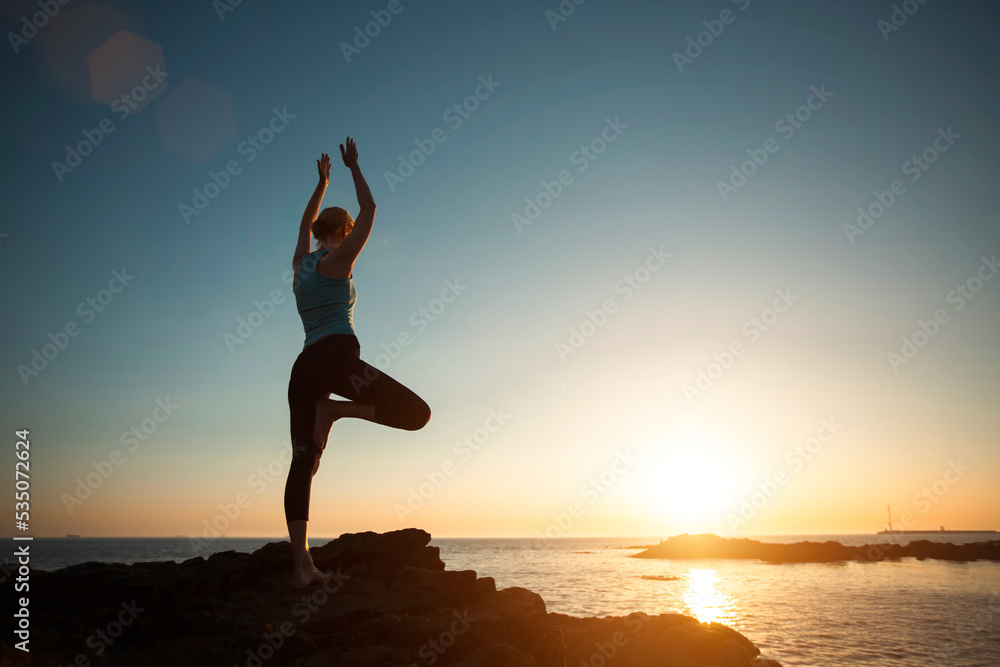 A woman does gymnastic yoga at sunset on the ocean.
