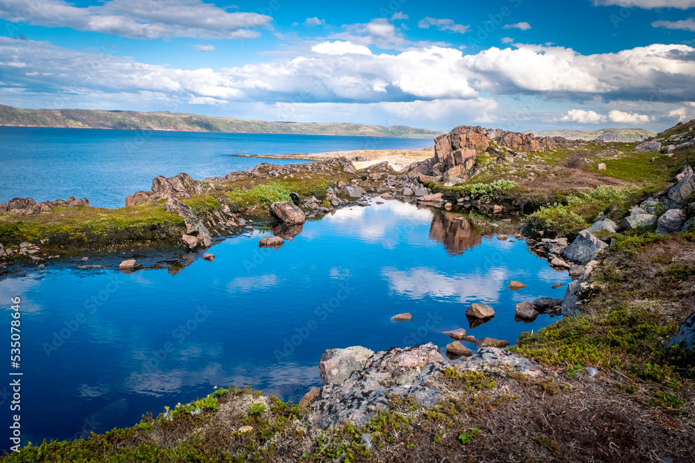 Natural pool in the rock. Blue sky with clouds reflected in clear water. Rocky coastline of Barents Sea near Teriberka. Scenery of Russian North. Kola Peninsula, Murmansk Oblast, Russia