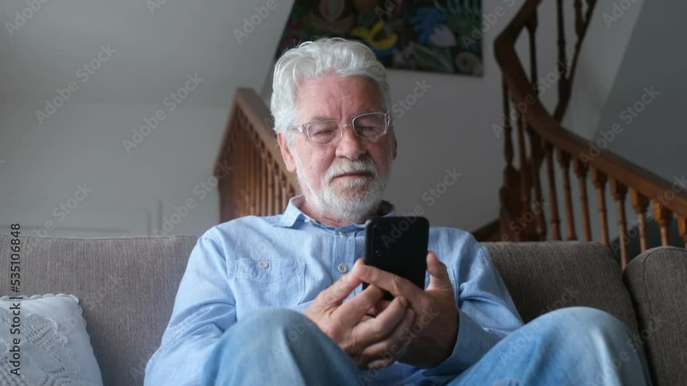 One old man using phone at home sitting on the sofa smiling and having fun learning chatting with friends and surfing on the web. Satisfied male senior indoor with smartphone. One old man using phone at home sitting on the sofa smiling and having fun learning chatting with friends and surfing on the web. Satisfied male senior indoor with smartphone.