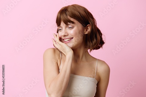 a modest, shy woman stands on a pink background and smiles pleasantly with her eyes closed and holding her hand near her head