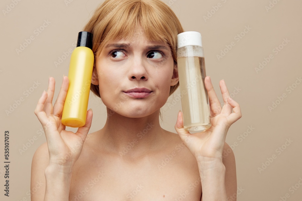 Fototapeta premium a funny, anxious woman stands on a beige background holding jars of care cosmetics in her hands and making an emotional face presses them to her head. Horizontal photo with empty space